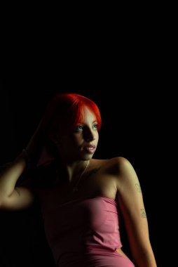 Güzel, genç, kızıl saçlı bir kadının portresi, oturan, pembe elbiseli, koyu arkaplanda fotoğraf için poz veren. Stüdyoda yapılmış bir portre.