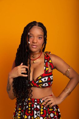  Saçları örgülü, geleneksel kıyafetler giyen, kameraya poz veren güzel bir genç kadın. Hardal renkli arka planda izole edilmiş. Kendine güvenen ve sakin biri..