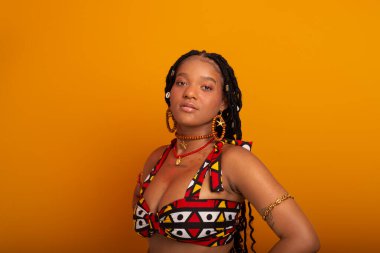  Saçları örgülü, geleneksel kıyafetler giyen, kameraya poz veren güzel bir genç kadın. Hardal renkli arka planda izole edilmiş. Kendine güvenen ve sakin biri..