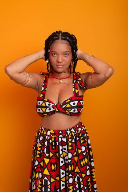 Saçında örgüleri olan, ayakta duran, geleneksel kıyafetler giyen, kameraya poz veren güzel bir genç kadın. Hardal renkli arka planda izole edilmiş. Kendine güvenen ve sakin biri..