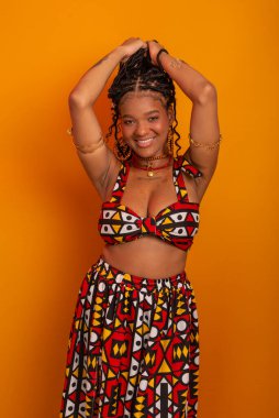 Saçında örgüleri olan, ayakta duran, geleneksel kıyafetler giyen, kameraya poz veren güzel bir genç kadın. Hardal renkli arka planda izole edilmiş. Kendine güvenen ve sakin biri..