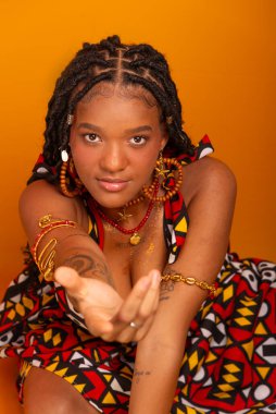 Saçında örgülerle oturan, geleneksel kıyafetler giyen, kameraya poz veren güzel bir genç kadın. Hardal renkli arka planda izole edilmiş. Kendine güvenen ve sakin biri..