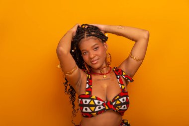 Saçında örgülerle oturan, geleneksel kıyafetler giyen, kameraya poz veren güzel bir genç kadın. Hardal renkli arka planda izole edilmiş. Kendine güvenen ve sakin biri..