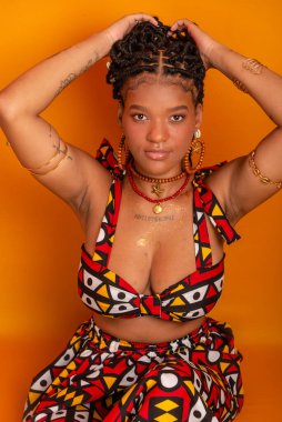 Saçında örgülerle oturan, geleneksel kıyafetler giyen, kameraya poz veren güzel bir genç kadın. Hardal renkli arka planda izole edilmiş. Kendine güvenen ve sakin biri..