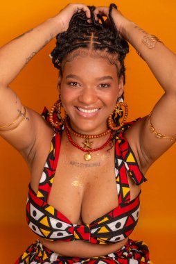 Saçında örgülerle oturan, geleneksel kıyafetler giyen, kameraya poz veren güzel bir genç kadın. Hardal renkli arka planda izole edilmiş. Kendine güvenen ve sakin biri..