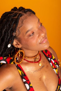 Saçları örgülü, geleneksel kıyafetler giyen ve kameraya poz veren güzel bir kadının dikey yakın plan portresi. Hardal renkli arka planda izole edilmiş. Kendine güvenen ve sakin biri..