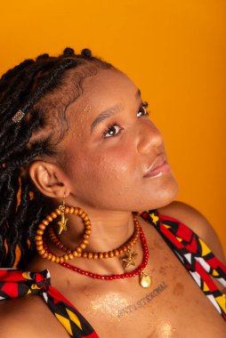 Saçları örgülü, geleneksel kıyafetler giyen ve kameraya poz veren güzel bir kadının dikey yakın plan portresi. Hardal renkli arka planda izole edilmiş. Kendine güvenen ve sakin biri..