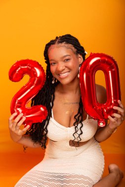 Beyaz elbiseli güzel bir doğum günü kızı turuncu renkli stüdyo zemininde oturuyor. Mutlu ve kendinden emin kişi.