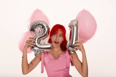 Pembe elbiseli genç kızıl saçlı kız elinde balonlarla iki numara ve yüz hizasında bir numara tutuyor. Beyaz arka planda izole edilmiş. Stüdyo çekimi