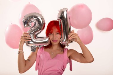 Pembe elbiseli genç kızıl saçlı kız elinde balonlarla iki numara ve yüz hizasında bir numara tutuyor. Beyaz arka planda izole edilmiş. Stüdyo çekimi