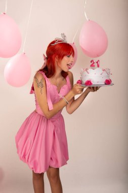 Kızıl saçlı genç bir kızın portresi, pembe elbiseli, elinde pasta, arkasında da hava balonları. Beyaz arka planda izole edilmiş. Stüdyo fotoğrafı