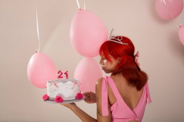 Elinde pasta tutan pembe elbiseli güzel kızıl saçlı doğum günü kızının portresi. Hava balonları arkada. Işık arka planında izole edilmiş. Stüdyo fotoğrafı