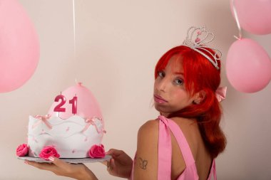 Elinde pasta tutan pembe elbiseli güzel kızıl saçlı doğum günü kızının portresi. Hava balonları arkada. Işık arka planında izole edilmiş. Stüdyo fotoğrafı