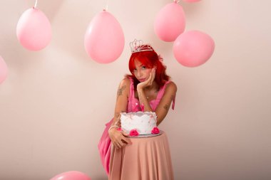 Doğum günü pastasının yanında çenesi açık pembe elbiseli genç ve güzel bir kız. Hava balonları arkada. Işık arka planında izole edilmiş. Stüdyo fotoğrafı