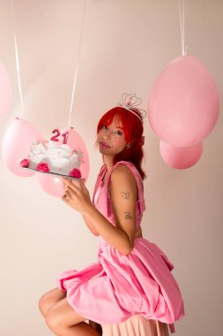 Genç, güzel, kızıl saçlı, pembe elbiseli, elinde doğum günü pastası olan, arka planda fotoğraf için poz veren bir kız. Hava balonları arkada. Stüdyo resmi