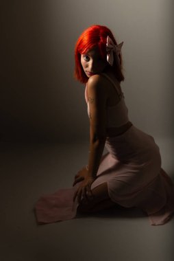 Pembe elbiseli genç, kızıl saçlı kadın yere çömelmiş, karanlık arka planda fotoğraf için poz veriyor. Stüdyo resmi