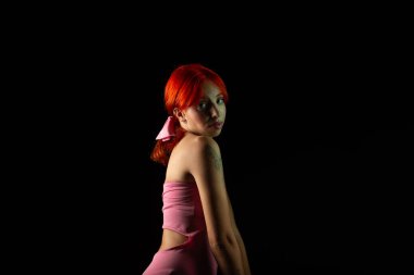 Güzel, genç, kızıl saçlı bir kadının portresi, oturan, pembe elbiseli, koyu arkaplanda fotoğraf için poz veren. Stüdyoda yapılmış bir portre.