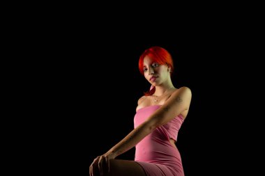 Güzel, genç, kızıl saçlı bir kadının portresi, oturan, pembe elbiseli, koyu arkaplanda fotoğraf için poz veren. Stüdyo resmi.