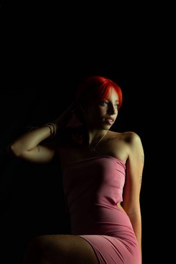 Güzel, genç, kızıl saçlı bir kadının portresi, oturan, pembe elbiseli, koyu arkaplanda fotoğraf için poz veren. Stüdyo resmi.