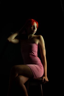 Güzel, genç, kızıl saçlı bir kadının portresi, oturan, pembe elbiseli, koyu arkaplanda fotoğraf için poz veren. Stüdyo resmi.