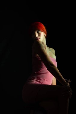 Güzel, genç, kızıl saçlı bir kadının portresi, oturan, pembe elbiseli, koyu arkaplanda fotoğraf için poz veren. Stüdyo resmi.
