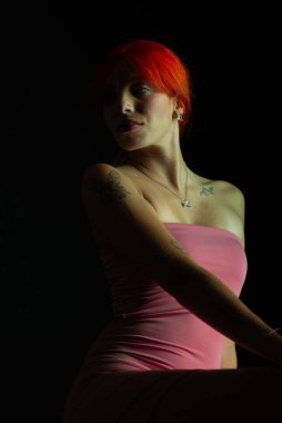 Güzel, genç, kızıl saçlı bir kadının portresi, oturan, pembe elbiseli, koyu arkaplanda fotoğraf için poz veren. Stüdyo resmi.