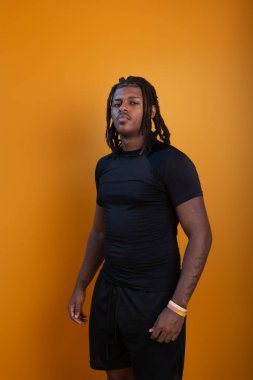 Şort giyen yakışıklı bir adam ve turuncu arka planda fotoğraf için poz veren siyah gömlekli. Kendine güvenen ve sakin biri..