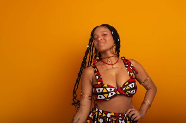  Saçları örgülü, geleneksel kıyafetler giyen, kameraya poz veren güzel bir genç kadın. Hardal renkli arka planda izole edilmiş. Kendine güvenen ve sakin biri..