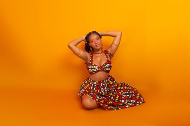 Saçında örgülerle oturan, geleneksel kıyafetler giyen, kameraya poz veren güzel bir genç kadın. Hardal renkli arka planda izole edilmiş. Kendine güvenen ve sakin biri..