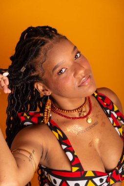 Saçları örgülü, geleneksel kıyafetler giyen ve kameraya poz veren güzel bir kadının dikey yakın plan portresi. Hardal renkli arka planda izole edilmiş. Kendine güvenen ve sakin biri..