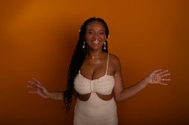 Beyaz elbiseli, mutlu ve parti konfetileri arasında gülümseyen genç bir doğum günü kızı. Koyu turuncu arka planda izole edilmiş. Mutlu ve kendinden emin kişi.