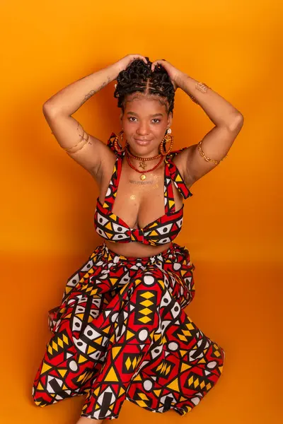 Saçında örgülerle oturan, geleneksel kıyafetler giyen, kameraya poz veren güzel bir genç kadın. Hardal renkli arka planda izole edilmiş. Kendine güvenen ve sakin biri..