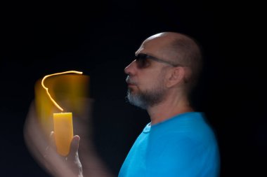 Mavi gömlekli kel adamın sanatsal portresi yanan sarı mumla hareket bulanıklığı yaratıyor. Hafif patika. Siyah arkaplan.