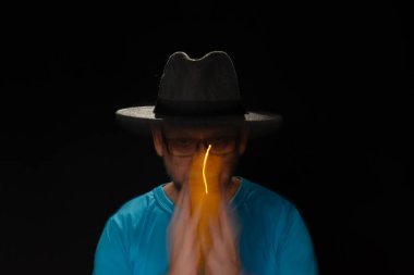 Mavi tişörtlü, sakallı, kel, şapkalı, yanan mumla ışık saçan adamın sanatsal portresi. Hareket bulanıklığı. Siyah arkaplan.