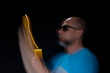 Mavi gömlekli kel adamın sanatsal portresi yanan sarı mumla hareket bulanıklığı yaratıyor. Hafif patika. Siyah arkaplan.