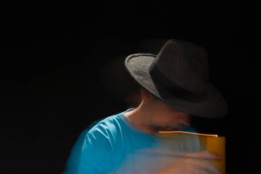 Mavi tişörtlü, sakallı, kel, şapkalı, yanan mumla ışık saçan adamın sanatsal portresi. Hareket bulanıklığı. Siyah arkaplan.