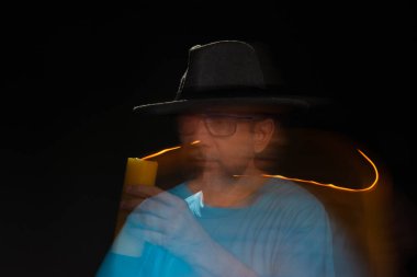 Mavi tişörtlü, sakallı, kel, şapkalı, yanan mumla ışık saçan adamın sanatsal portresi. Hareket bulanıklığı. Siyah arkaplan.