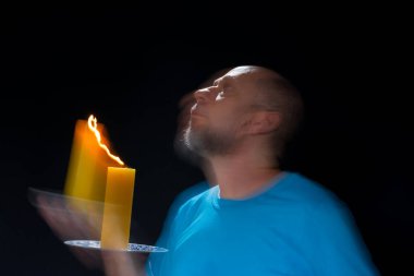 Mavi gömlekli kel adamın sanatsal portresi yanan sarı mumla hareket bulanıklığı yaratıyor. Hafif patika. Siyah arkaplan.
