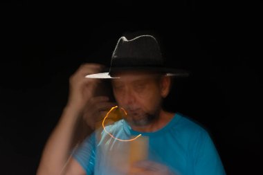 Mavi tişörtlü, sakallı, kel, şapkalı, yanan mumla ışık saçan adamın sanatsal portresi. Hareket bulanıklığı. Siyah arkaplan.