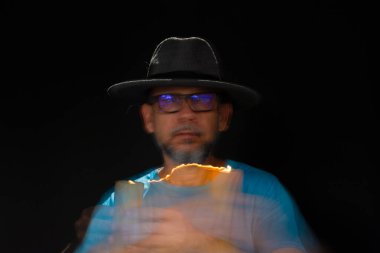 Mavi tişörtlü, sakallı, kel, şapkalı, yanan mumla ışık saçan adamın sanatsal portresi. Hareket bulanıklığı. Siyah arkaplan.