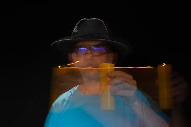 Mavi tişörtlü, sakallı, kel, şapkalı, yanan mumla ışık saçan adamın sanatsal portresi. Hareket bulanıklığı. Siyah arkaplan.