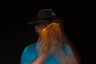 Mavi tişörtlü, sakallı, kel, şapkalı, yanan mumla ışık saçan adamın sanatsal portresi. Hareket bulanıklığı. Siyah arkaplan.