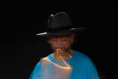 Mavi tişörtlü, sakallı, kel, şapkalı, yanan mumla ışık saçan adamın sanatsal portresi. Hareket bulanıklığı. Siyah arkaplan.