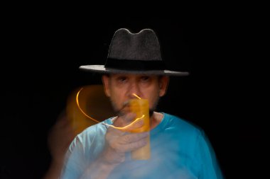 Mavi tişörtlü, sakallı, kel, şapkalı, yanan mumla ışık saçan adamın sanatsal portresi. Hareket bulanıklığı. Siyah arkaplan.