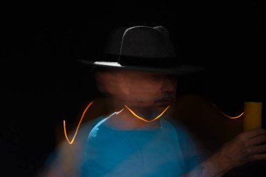 Mavi tişörtlü, sakallı, kel, şapkalı, yanan mumla ışık saçan adamın sanatsal portresi. Hareket bulanıklığı. Siyah arkaplan.