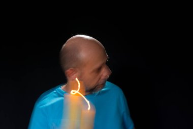 Mavi gömlekli kel adamın sanatsal portresi yanan sarı mumla hareket bulanıklığı yaratıyor. Hafif patika. Siyah arkaplan.