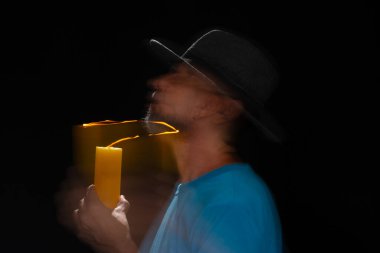 Mavi tişörtlü, sakallı, kel, şapkalı, yanan mumla ışık saçan adamın sanatsal portresi. Hareket bulanıklığı. Siyah arkaplan.