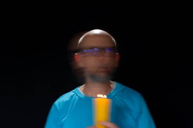 Mavi gömlekli kel adamın sanatsal portresi yanan sarı mumla hareket bulanıklığı yaratıyor. Hafif patika. Siyah arkaplan.