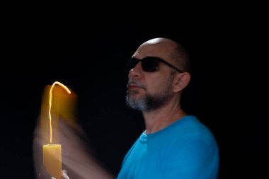 Mavi gömlekli kel adamın sanatsal portresi yanan sarı mumla hareket bulanıklığı yaratıyor. Hafif patika. Siyah arkaplan.