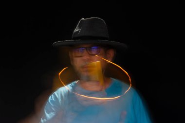 Mavi tişörtlü, sakallı, kel, şapkalı, yanan mumla ışık saçan adamın sanatsal portresi. Hareket bulanıklığı. Siyah arkaplan.